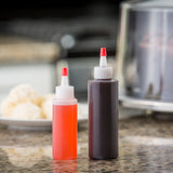 Two TableCraft squeeze bottles with dishwasher safe, cone top caps hold red and dark brown liquids on a kitchen counter. In the blurred background, there’s a plate with ice cream scoops and a stainless steel appliance.