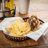 A brown Chicago Basket lined with white paper holds a pile of ridged potato chips and a sandwich filled with shredded beef, topped with chopped peppers and garlic. Salt and pepper shakers and a cup are blurred in the background on a wooden table.