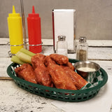 A green plastic, heat resistant basket filled with buffalo chicken wings, celery sticks, and a metal cup of dipping sauce sits on a white wooden table. In the background are yellow and red squeeze bottles, a napkin holder, and salt and pepper shakers.