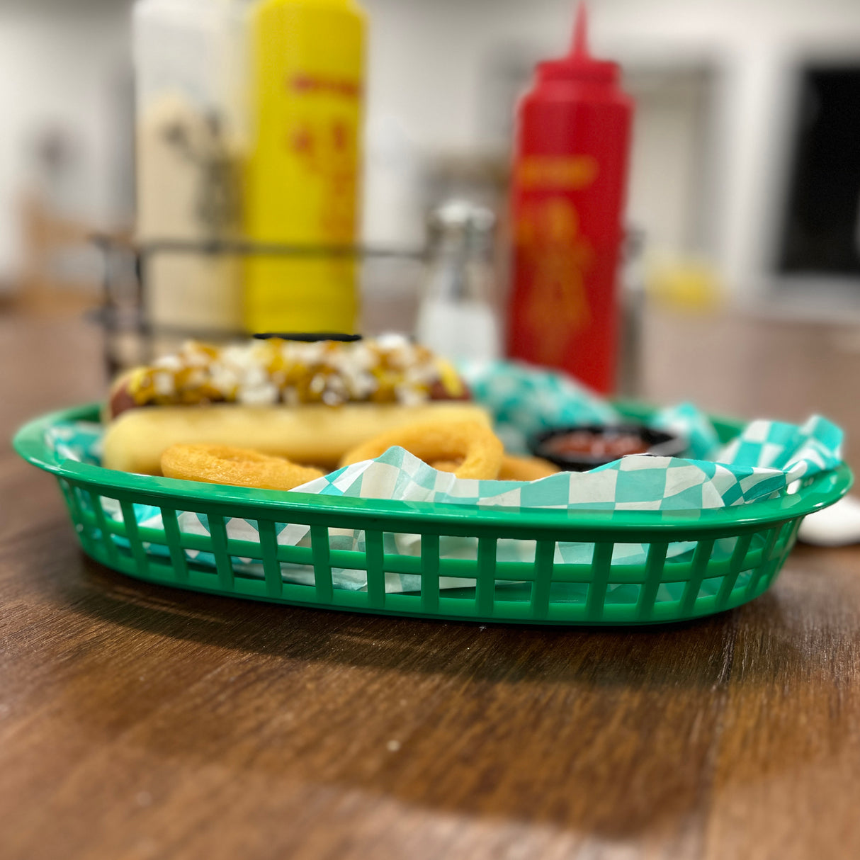 A green plastic Chicago basket lined with blue-and-white checkered paper holds a hot dog topped with relish, onions, and mustard, alongside onion rings. In the blurred background are ketchup and mustard bottles on a wooden table.
