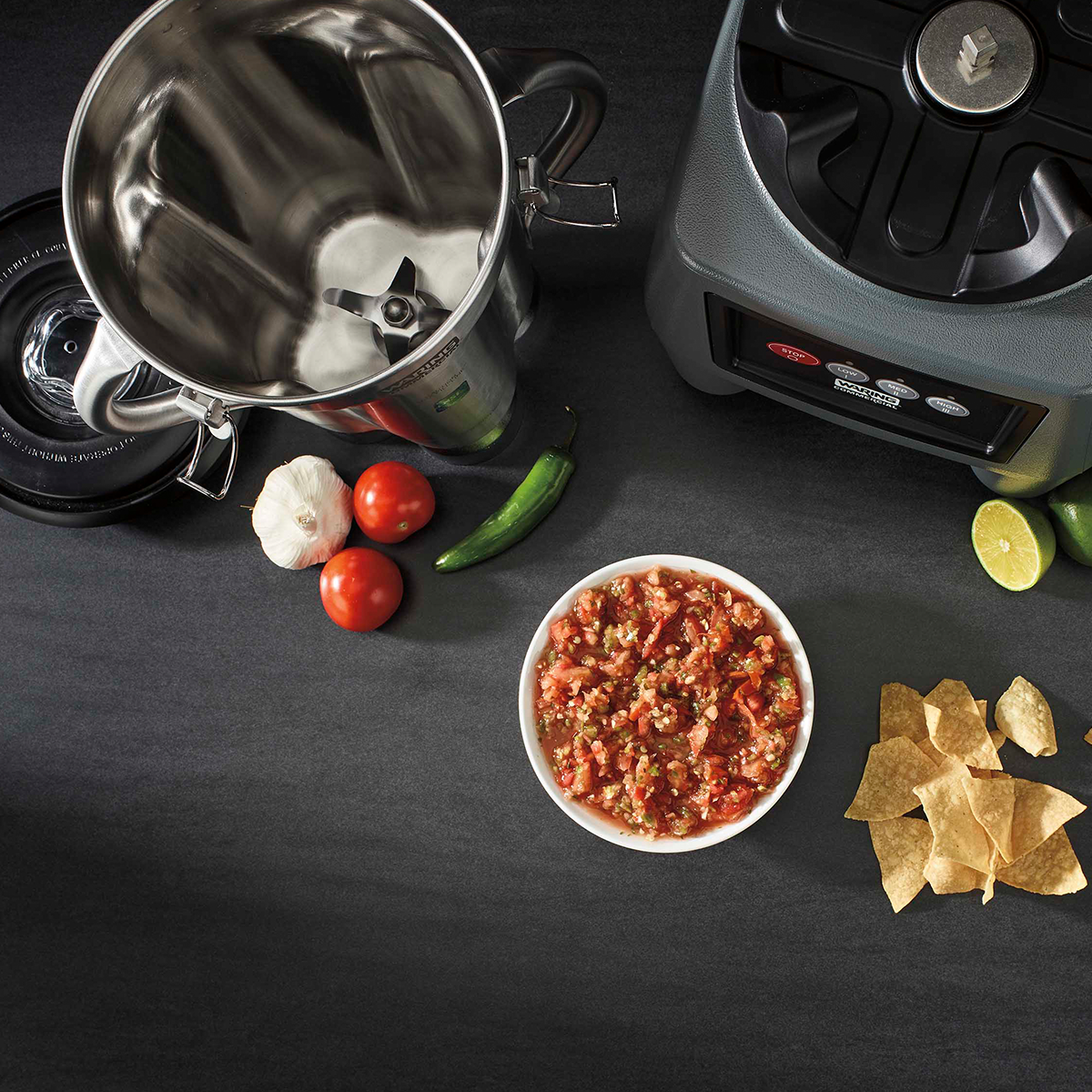 Overhead view of a Commercial Blender with a stainless steel container, blender lid, tomatoes, garlic, serrano pepper, lime, a bowl of chunky salsa, and tortilla chips on a dark countertop. The 4 liter blender is open and empty.