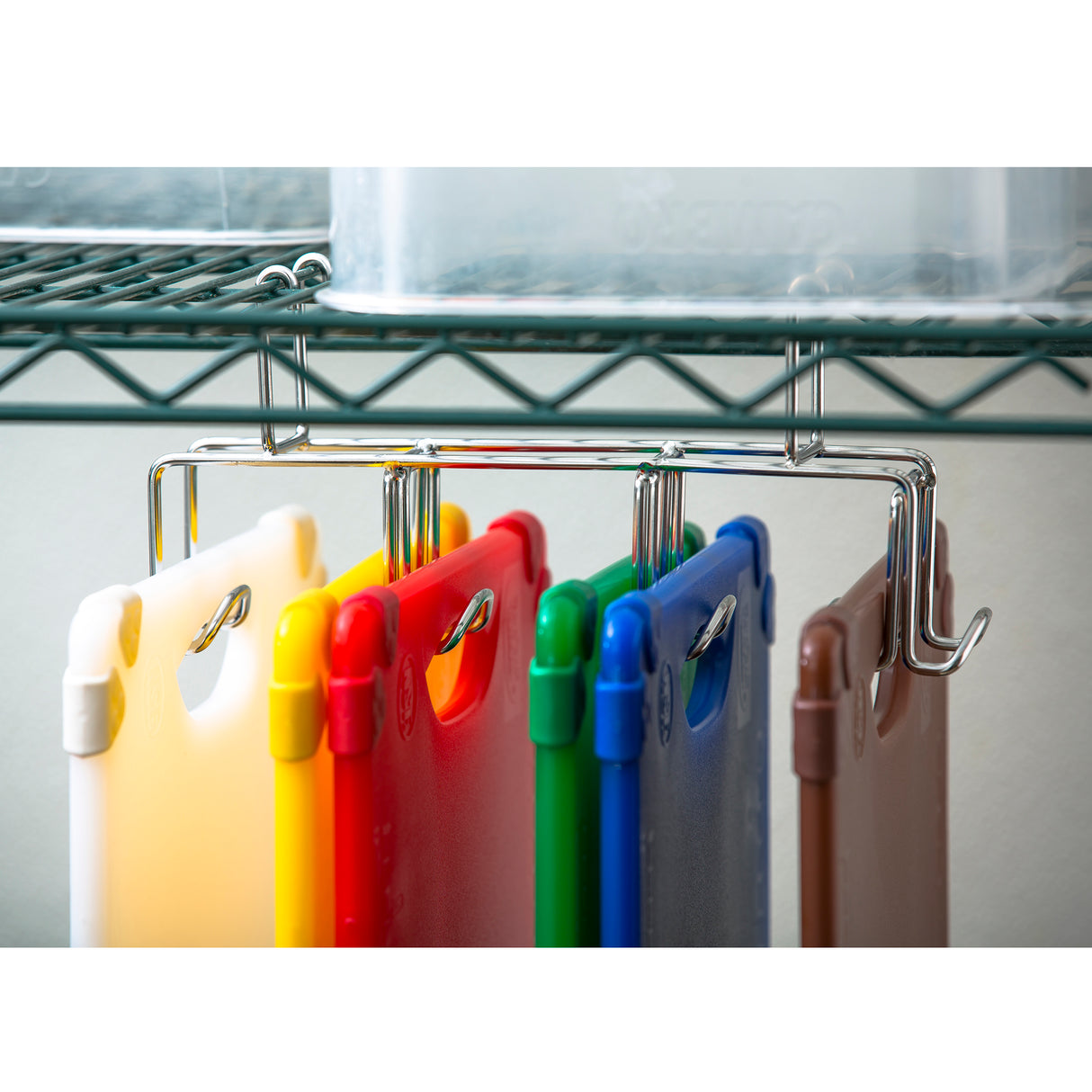 Six grippy cutting boards in white, yellow, red, green, blue, and brown hang vertically on metal hooks attached to a chrome wire shelf. A clear plastic container sits above them. The background is neutral and out of focus. All boards are dishwasher safe.