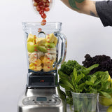 A person adds red grapes to a Breville Commercial Super Q Pro Blender filled with sliced bananas, green apples, and pineapple. Fresh greens like kale and mint sit nearby. The persons tattooed arm is visible by the 68 oz capacity stainless steel base.
