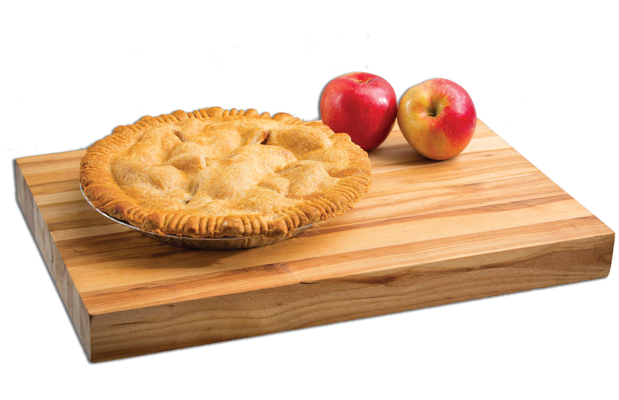 A golden-brown apple pie with a crimped crust sits on an edge-grain butcher cutting board next to two red apples. The white background highlights the pie and apples as the focal point.