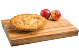 A golden-brown apple pie with a crimped crust sits on an edge-grain butcher cutting board next to two red apples. The white background highlights the pie and apples as the focal point.