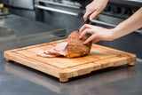 A person slices a cooked, glazed ham on a non-slip carving board in a commercial kitchen. The ham’s browned outer layer is being cut into even slices with a sharp knife, surrounded by stainless steel counters and kitchen equipment.