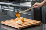 A hand uses a ladle to pour thick, orange soup into a hollowed-out round bread loaf on a non-slip wood carving board, with the bread top set aside. The background reveals a stainless steel kitchen with stoves and counters.