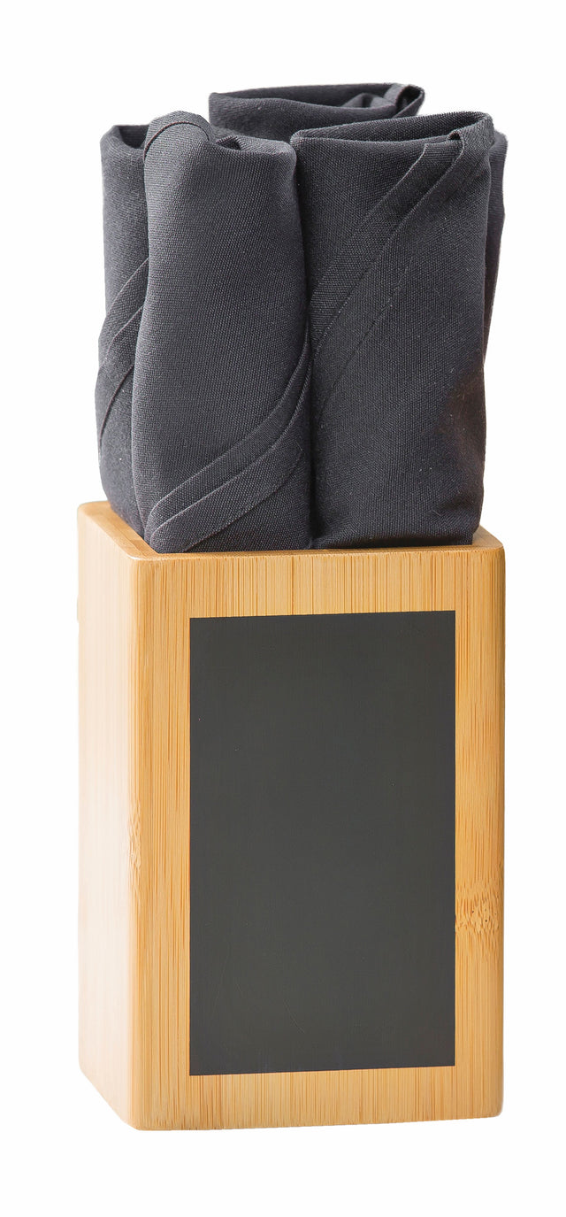 A wooden TableCraft Products bamboo cutlery holder with a blank black write-on label holds three neatly rolled, dark gray cloth napkins. The smooth block contrasts the rosebud-shaped napkins against its light wood for a striking display.