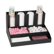A black condiment organizer holds paper cups, plastic lids, pink and white sweetener packets, dairy creamer packets, white stir sticks, and red straws, neatly separated in compartments for convenient coffee or beverage preparation.