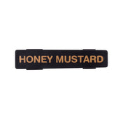 A rectangular black plastic TableCraft name tag with gold capital letters reading “HONEY MUSTARD.” The sign has two small clips on the back for attachment, and the background is white.