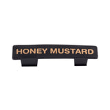 A black rectangular Name Tag label with gold capital letters spelling HONEY MUSTARD. Featuring two small tabs on the bottom for easy attachment to a dispenser or container, this tag is dishwasher safe. The background is plain white.