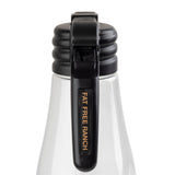 A close-up of a clear TableCraft bottle with a black flip-top cap. The cap features a vertical name tag that reads “FAT FREE RANCH” in gold letters on black. The white liquid inside suggests it contains fat free ranch salad dressing.