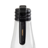 A clear bottle with a black polypropylene flip-top lid is shown close-up. The lid has a ribbed design and a black lever with RANCH written in gold letters like a Ranch name tag. The background is white, keeping the focus on the bottle and lid.