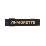 A black rectangular TableCraft name tag features the word VINAIGRETTE in bold, uppercase, gold letters centered on it. The label has small, raised tabs on its top and bottom edges against a plain white background.