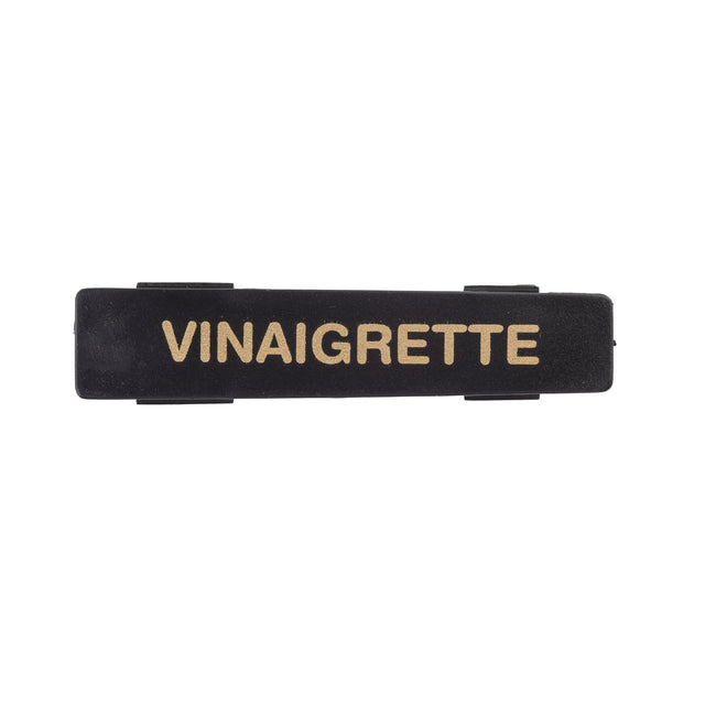 A black rectangular TableCraft name tag features the word VINAIGRETTE in bold, uppercase, gold letters centered on it. The label has small, raised tabs on its top and bottom edges against a plain white background.