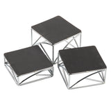 Three modern square display stands with black tops and chrome plated silver metal frames are arranged at different heights. The geometric, angular frames create a striking look as the riser set is grouped closely on a white background.