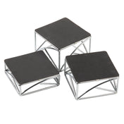 Three modern square display stands with black tops and chrome plated silver metal frames are arranged at different heights. The geometric, angular frames create a striking look as the riser set is grouped closely on a white background.