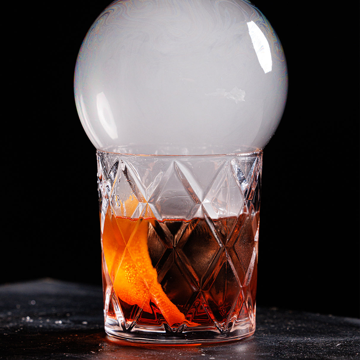 A crystal glass with an orange peel and dark cocktail sits on a dark surface. Using a Smoke Bubble Kit, a large, transparent smoke bubble encloses aromatic smoke over the rim, creating a dramatic and elegant presentation against a black background.
