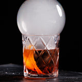 A crystal glass with an orange peel and dark cocktail sits on a dark surface. Using a Smoke Bubble Kit, a large, transparent smoke bubble encloses aromatic smoke over the rim, creating a dramatic and elegant presentation against a black background.