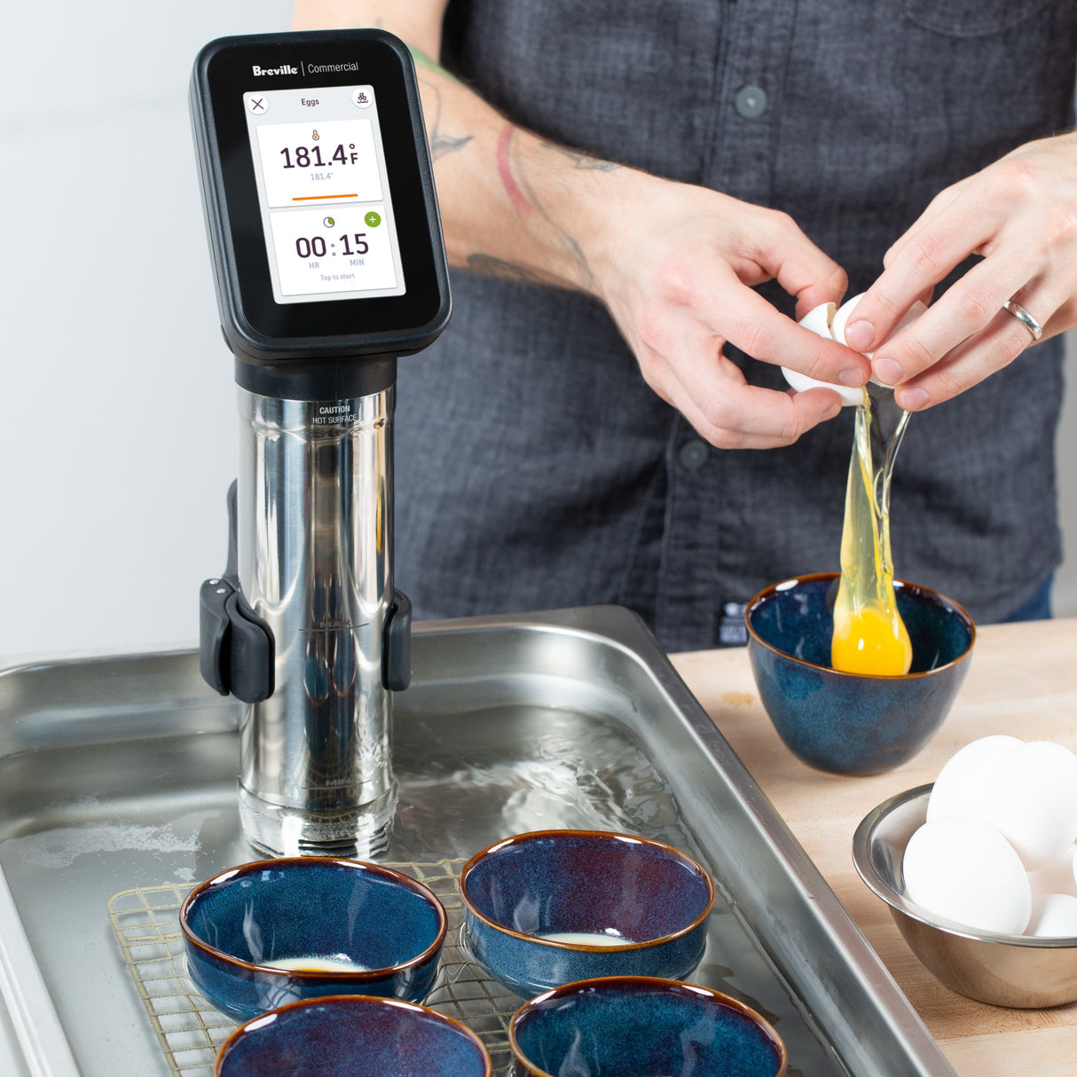 A person cracks a raw egg into a blue bowl next to a Breville Commercial Sous Vide Immersion Circulator set to 181.4°F for 15 minutes; four empty bowls and a tray sit in the water bath, with extra eggs on the light wooden surface.