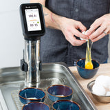 A person cracks a raw egg into a blue bowl next to a Breville Commercial Sous Vide Immersion Circulator set to 181.4°F for 15 minutes; four empty bowls and a tray sit in the water bath, with extra eggs on the light wooden surface.