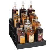 A black polystyrene bottle organizer holds twelve bottles of flavored syrups, each with pump dispensers and labeled with flavors like vanilla, caramel, hazelnut, and almond. The tiered rack sits neatly on the countertop for easy access.