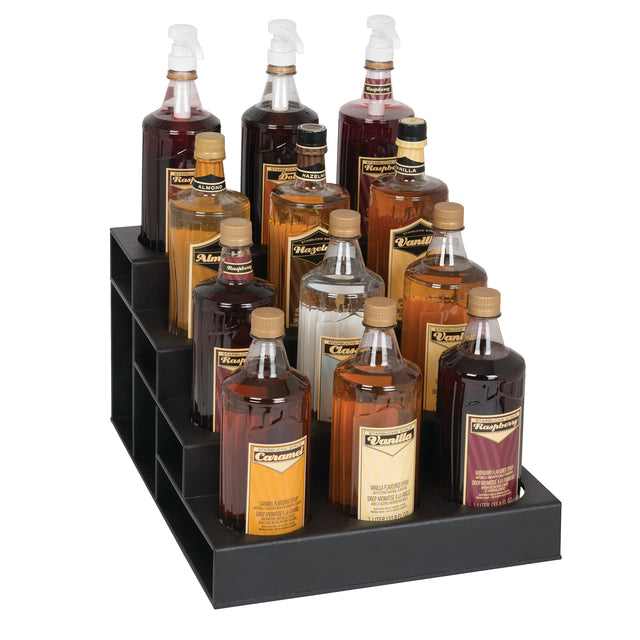 A black polystyrene bottle organizer holds twelve bottles of flavored syrups, each with pump dispensers and labeled with flavors like vanilla, caramel, hazelnut, and almond. The tiered rack sits neatly on the countertop for easy access.