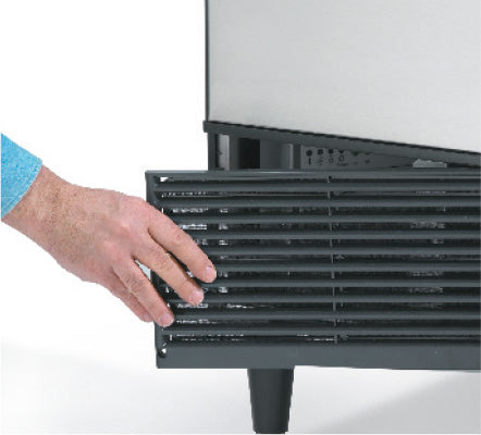 A person’s hand is removing the black vent cover from the bottom front panel of an undercounter ice maker, exposing the coils or internal components behind the vent. The appliance stands on small legs.