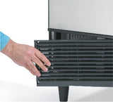 A person’s hand is pulling open a black vented grille at the bottom front of an undercounter ice maker, likely a cube style ice maker or Scotsman ice maker, which has a metallic finish and stands on small legs. The person wears a long-sleeved blue shirt.