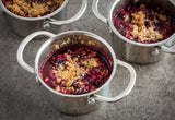 Three small stainless steel pots filled with berry crumble sit on a gray, textured surface. The golden, crispy topping covers a vibrant baked berry filling. Each mini server has two handles and a rustic, homemade appearance.