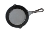 A round, black cast iron skillet with a textured surface, two small pouring spouts on the rim, and a long handle with a hole at the end. This induction ready skillet is shown from above on a white background.