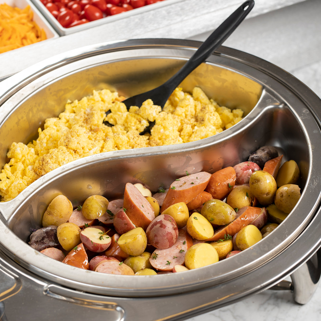 A stainless steel buffet dish contains scrambled eggs on one side and sliced sausages with small potatoes on the other. A black serving spoon rests in the eggs. In the background, trays of shredded cheese and cherry tomatoes are partially visible.