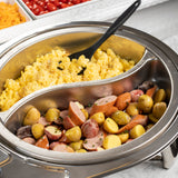A stainless steel buffet dish contains scrambled eggs on one side and sliced sausages with small potatoes on the other. A black serving spoon rests in the eggs. In the background, trays of shredded cheese and cherry tomatoes are partially visible.
