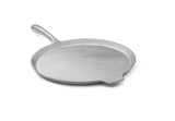 A flat, round, silver cast aluminum pizza pan with a slightly raised edge, a long curved handle on one side, and a small helper tab on the opposite side. The smooth surface appears against a plain white background.