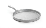 A metallic, silver-colored round cast aluminum frying pan with a textured surface and single handle angles upward, set on a plain white background. The empty pan is photographed from a slight angle, showing its depth and sturdy construction.
