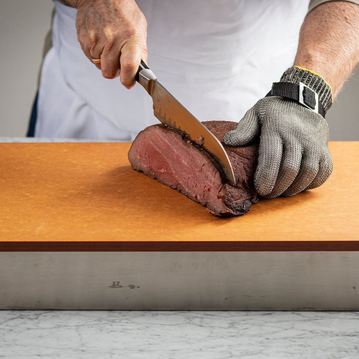 A person wearing a cut-resistant metal mesh glove slices rare roast beef on a Versa-Tile Carving Station with a large knife. Dressed in a white apron, they stand at a marble countertop, using this dishwasher safe carving station for easy cleanup.