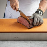 A person wearing a cut-resistant metal mesh glove slices rare roast beef on a Versa-Tile Carving Station with a large knife. Dressed in a white apron, they stand at a marble countertop, using this dishwasher safe carving station for easy cleanup.