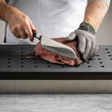 A person wearing a white apron and a cut-resistant glove slices cooked meat with a large knife on a TableCraft CW6432BK heat resistant carving station, featuring a perforated black cutting board atop a marble counter.