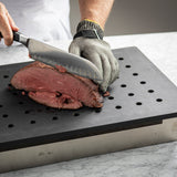 A person wearing a cut-resistant glove slices cooked beef on a TableCraft CW6432BK perforated cutting board atop a marble countertop, creating a heat resistant carving station while steadying the meat with one hand and slicing with a chef’s knife.