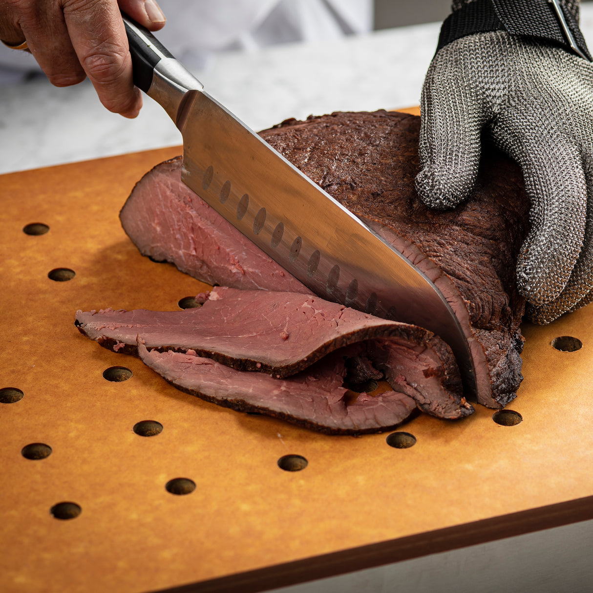 A person wearing a metal mesh glove slices roast beef on a heat resistant Versa-Tile Carving Station cutting board with evenly spaced holes. The beef is cooked rare, with a pink interior and dark brown crust.