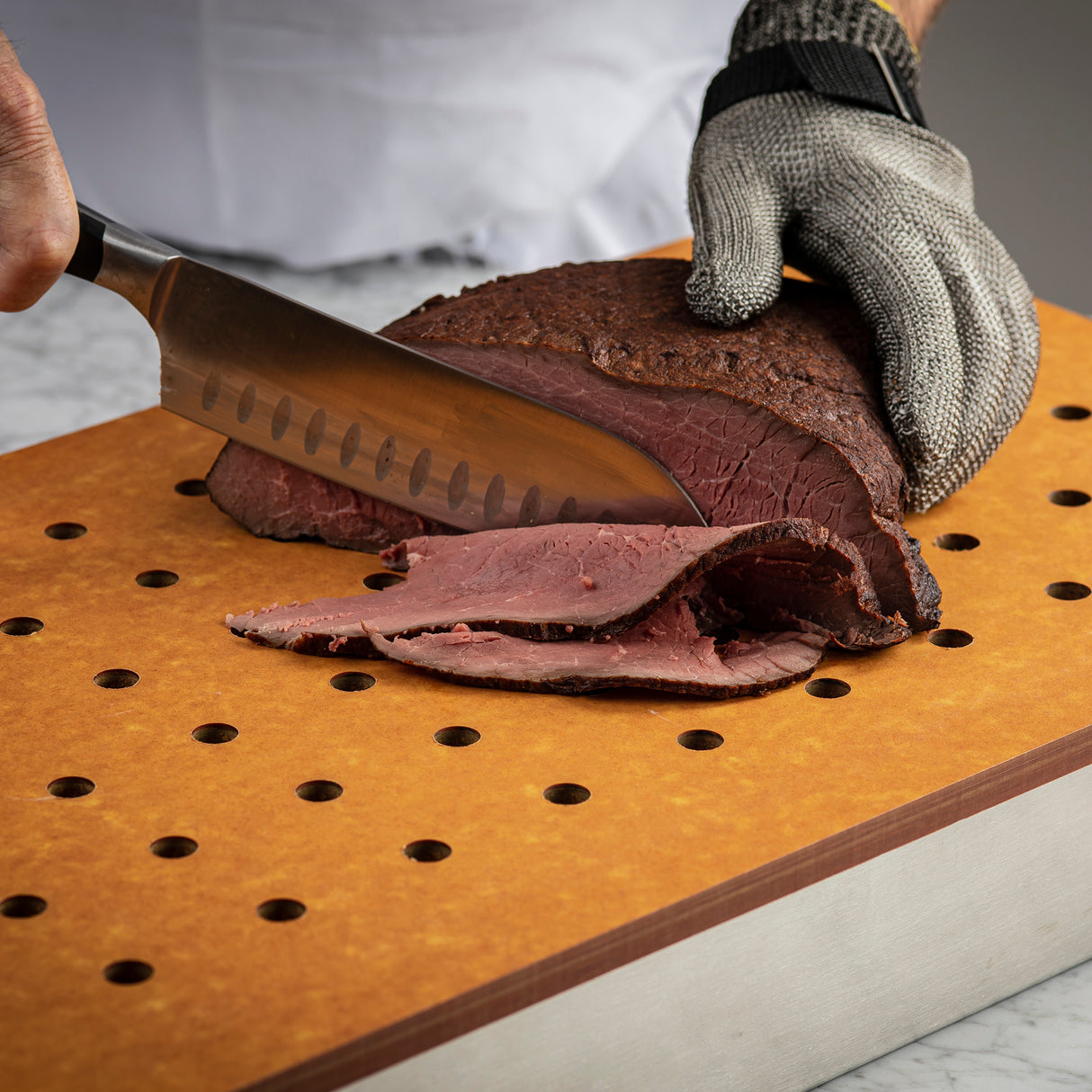 A person wearing a protective glove slices cooked roast beef on a heat resistant Versa-Tile Carving Station cutting board using a large knife. The yellow-brown, perforated board rests on a light-colored countertop.