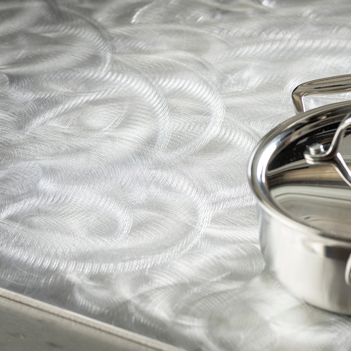 A shiny, dishwasher safe stainless steel pan with a lid sits on the edge of a metallic countertop, its brushed texture reflecting light and creating intricate, circular patterns across the surface.