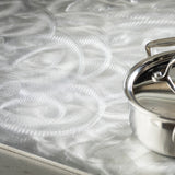 A shiny, dishwasher safe stainless steel pan with a lid sits on the edge of a metallic countertop, its brushed texture reflecting light and creating intricate, circular patterns across the surface.