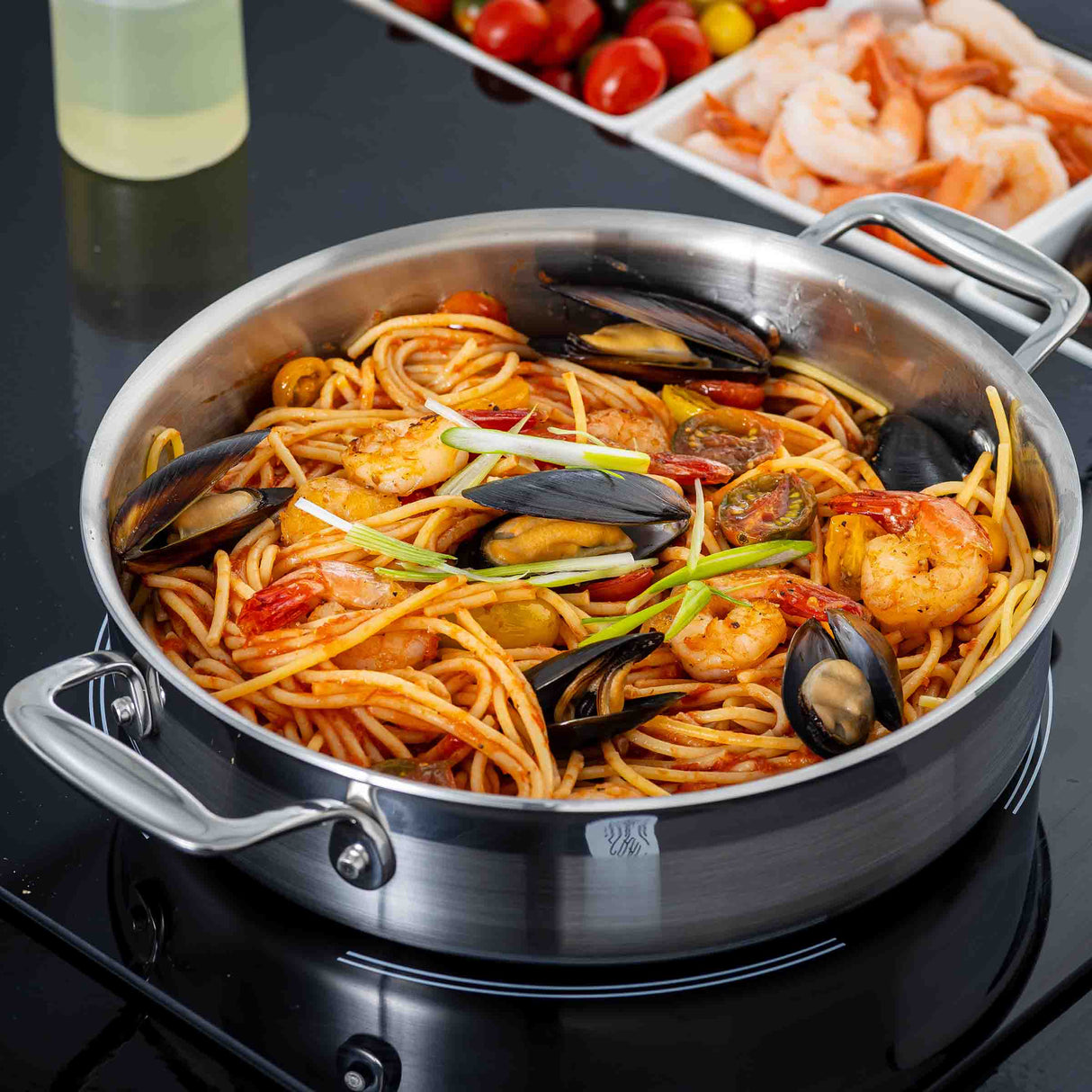 A tri-ply stainless steel Brazier Pan filled with spaghetti, shrimp, mussels, cherry tomatoes, and green onion garnish sits on a stovetop. In the background are raw shrimp, cherry tomatoes, and a bottle of oil.