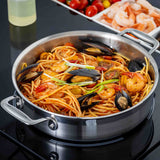 A tri-ply stainless steel Brazier Pan filled with spaghetti, shrimp, mussels, cherry tomatoes, and green onion garnish sits on a stovetop. In the background are raw shrimp, cherry tomatoes, and a bottle of oil.