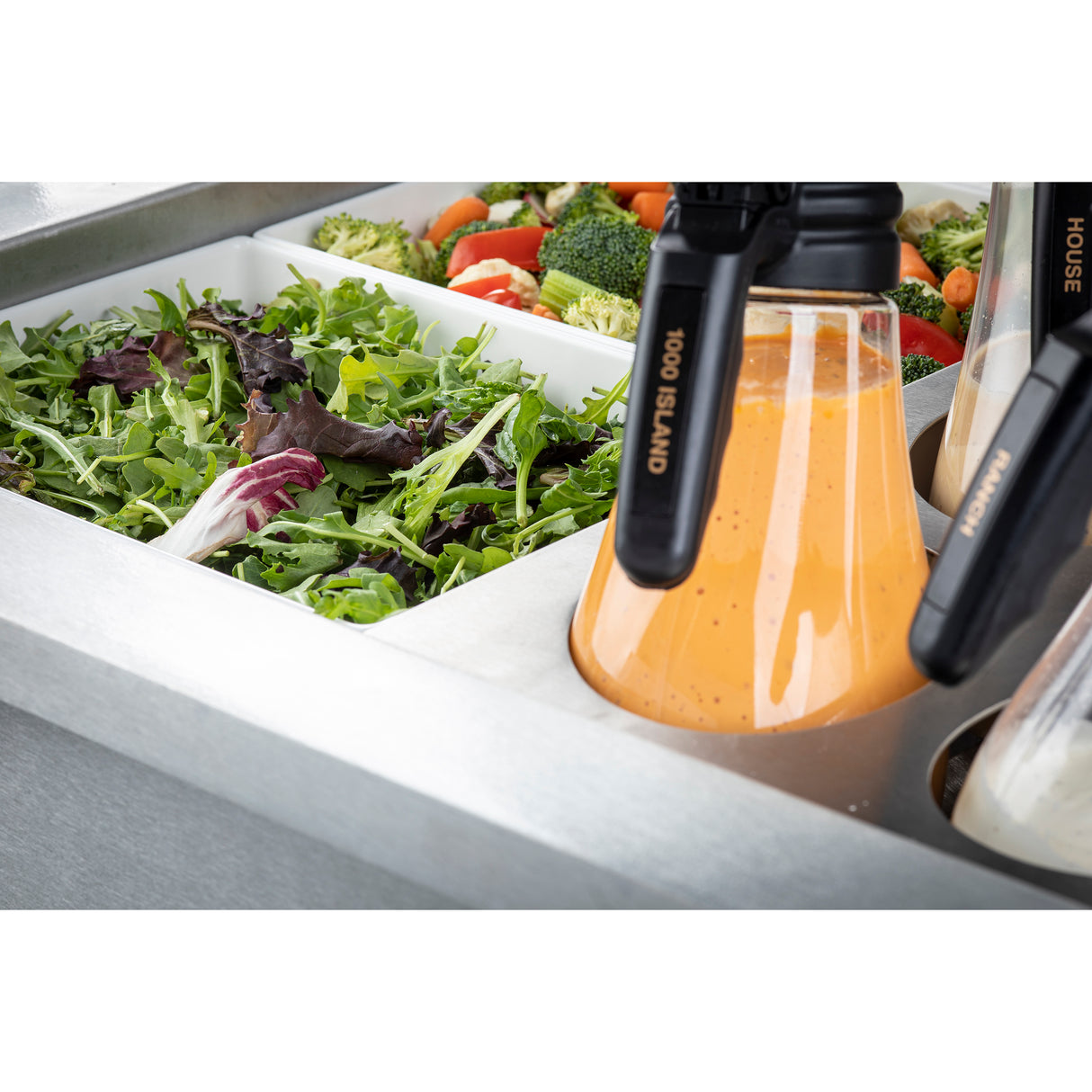A salad bar featuring a section of mixed leafy greens, chopped broccoli, and other vegetables is set beside a TableCraft CW8006BRA Double Ice Well, keeping large bottles of “1000 Island” and “House” dressings perfectly chilled.