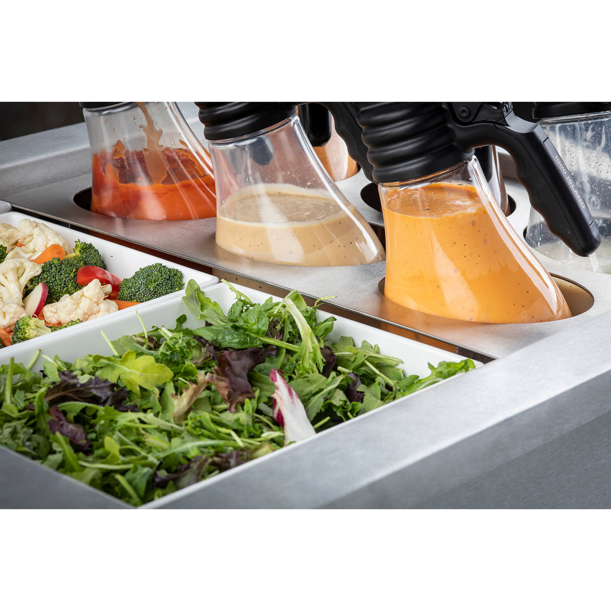 A salad bar with a tray of mixed greens in the foreground and containers of vegetable salad and various dressings in handled dispensers, all set in a stainless steel counter featuring a Double Ice Well for optimal freshness.