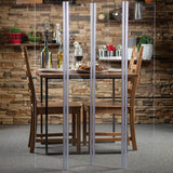 A dining table set for two with wooden chairs is divided by a clear acrylic safety shield. The table has wine glasses, plates, and condiments. A stone wall, shelves with plants, bottles, and kitchenware in the background create a cozy atmosphere.