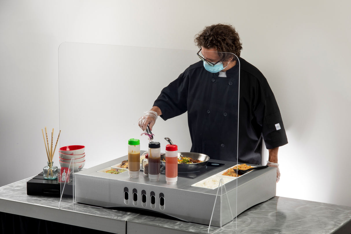 A chef in a black coat, gloves, and face mask cooks food in a pan behind a TableCraft acrylic shield. Various condiments, ingredients, and bowls are neatly arranged on the counter, creating a clean and organized action station.