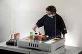 A chef in a black coat, gloves, and face mask cooks food in a pan behind a TableCraft acrylic shield. Various condiments, ingredients, and bowls are neatly arranged on the counter, creating a clean and organized action station.